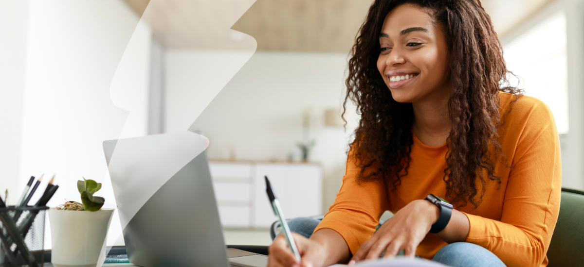 Black woman sitting at desk, using computer writing in notebook