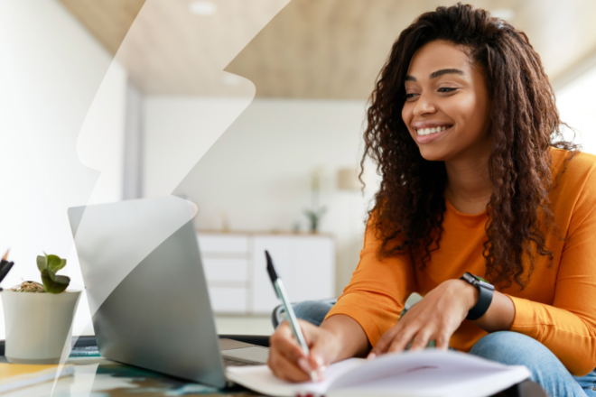 Black woman sitting at desk, using computer writing in notebook